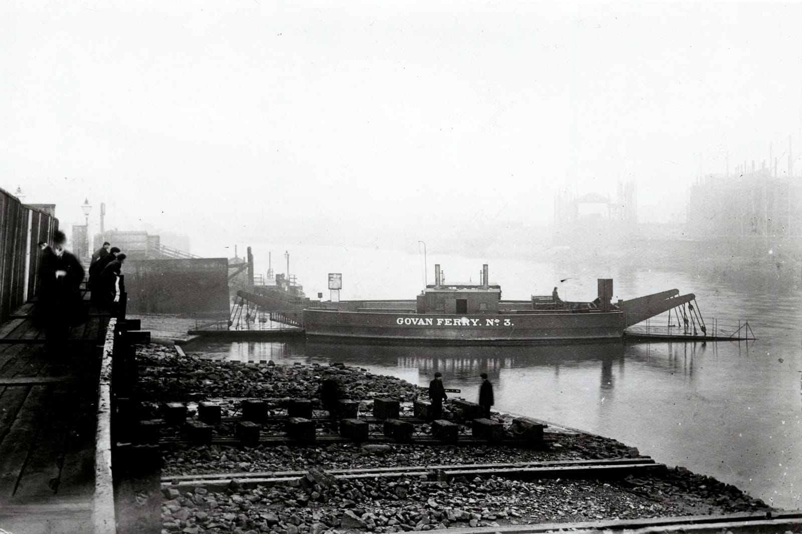 Black and white image of a pebbled dock, with a view of the Clydebank. A small boat is docked - in white lettering on its side, it reads 'Govan Ferry, no.3'