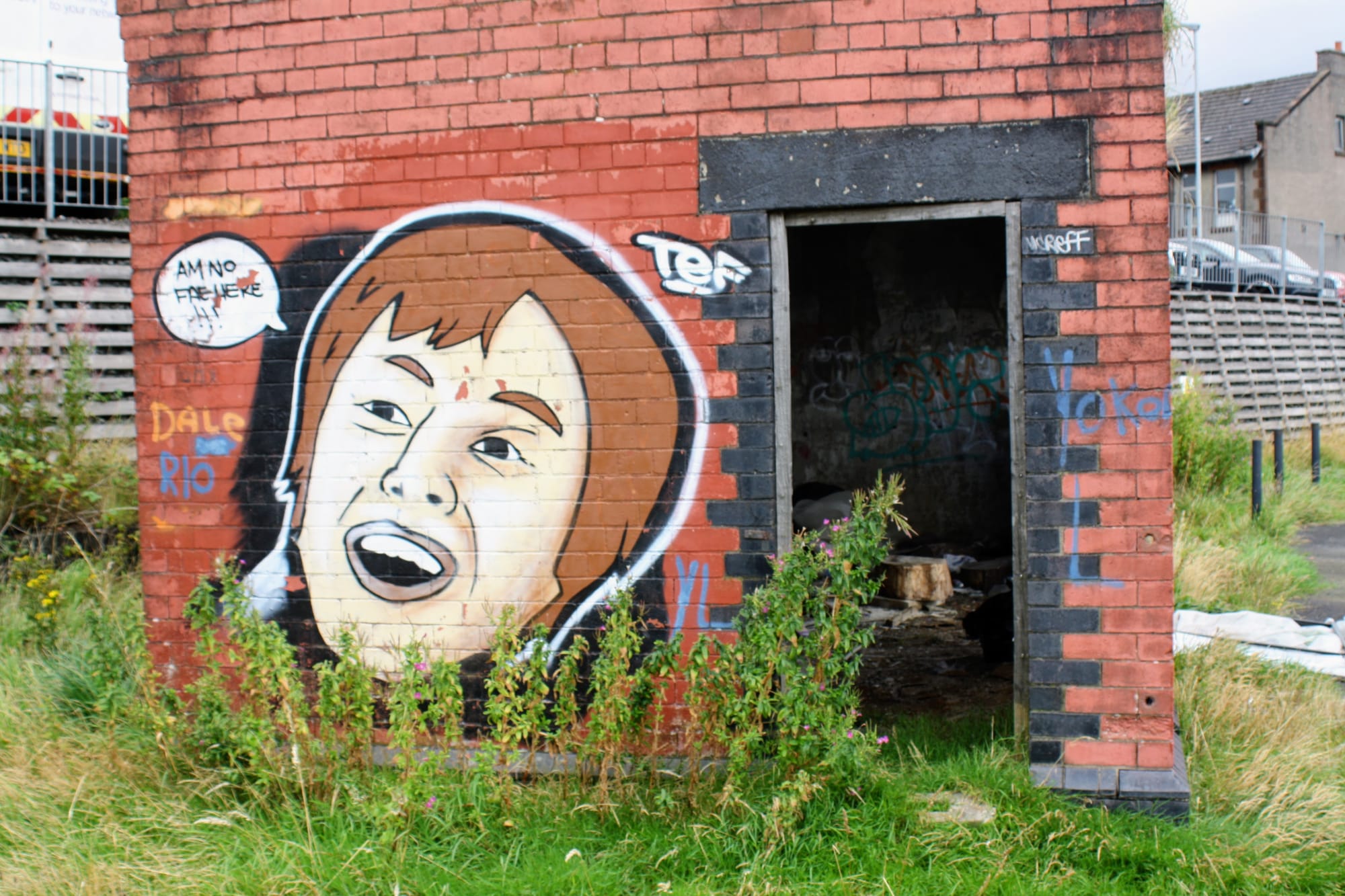 Image is of a red-brick bus shelter, surrounded by grass and a few weeds. On the right side of the bus shelter is a doorframe, picked out in black bricks. On the left side is a solid brick wall, painted with the mural of a brunette, Causcasian man's face. A speech bubble near the man's mouth reads 'Am no fae here'.