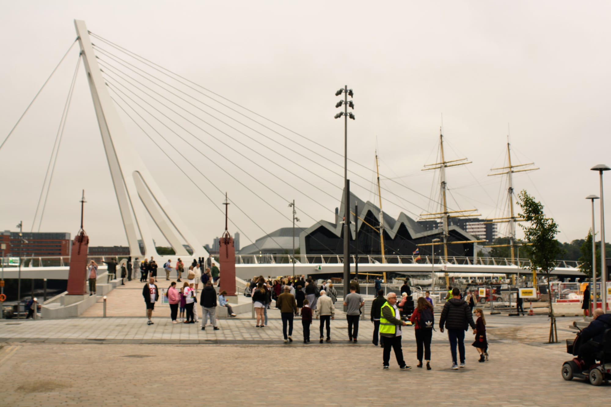 Image shows people milling on grey pavement at the September 2024 opening of Glasgow's new pedestrian bridge, connecting Govan and Partick. The bridge is large and white, with suspension cables jutting out from a prong that extends into the sky.