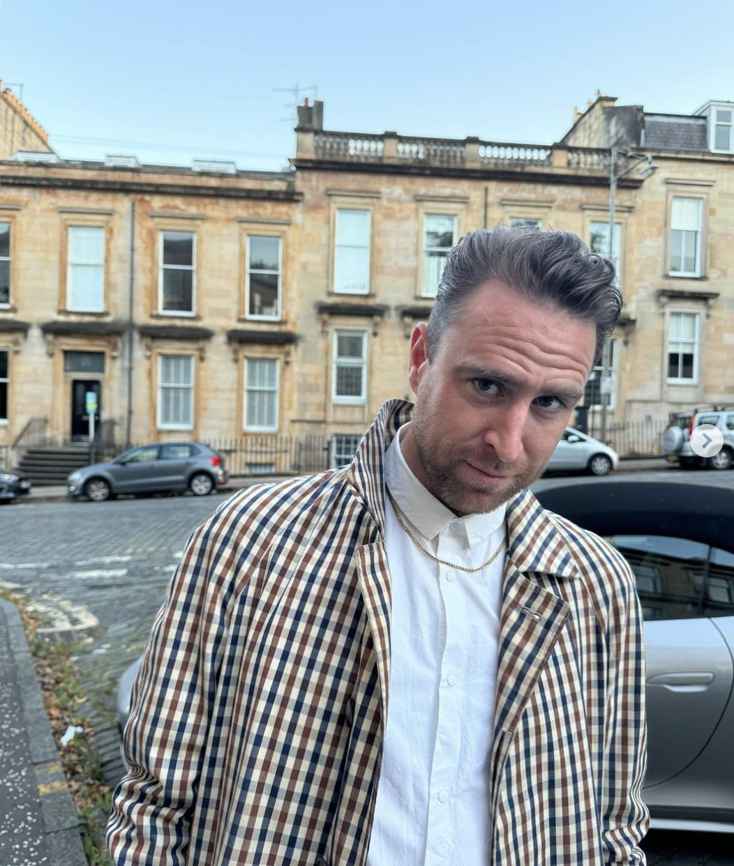 Image shows DJ Jack Revill, aka Jackmaster, who passed away on 12 October in Ibiza. Revill is looking at the camera and wearing a white collared shirt, a small gold chain and a checked jacket. In the background is a row of Glasgow Victorian buildings.